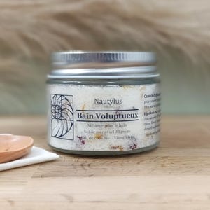 Op de afbeelding: Een glazen pot met een zilveren deksel gevuld met witte badzouten en gedroogde bloemen. Het etiket vermeldt "Nautylus Bain Voluptueux" en somt de ingrediënten op: "Mélange pour le bain, Sel de mer et sel d'Epsom, lait de coco bio, Ylang Ylang".