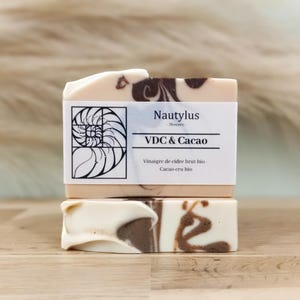 Puede incluir: Dos barras de jabón hechas a mano con un patrón de remolino en tonos marrones y blancos. La barra superior tiene una etiqueta que dice "Nautilus Skincare" y "VDC & Cacao" con el texto "Vinaigre de cidre brut bio - Cacao cru bio".