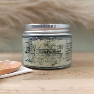 Può includere: Un barattolo di vetro con un coperchio argentato contenente un bagno schiumoso etichettato "Nautylus Bain Purifiant". L'etichetta elenca anche gli ingredienti: Sale marino, Sale di Epsom, Eucalipto e Tea Tree.
