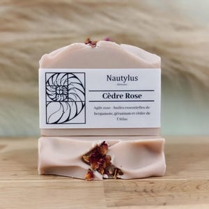 Puede incluir: Dos barras de jabón de color rosa pálido con pétalos de rosa secos. La barra superior tiene una etiqueta blanca con el texto "Nautylus - Skincare - Cèdre Rose Agile rose - huiles essentielles de bergamote, géranium et cèdre de l'Atlas".