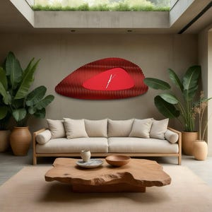 May include: A red, abstract wall clock with a white clock face and black hands hangs on a beige wall in a modern living room. The clock is surrounded by plants and a beige sofa with white cushions.