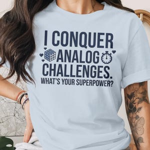 Pode incluir: T-shirt azul claro com o texto "I CONQUER ANALOG CHALLENGES. WHAT'S YOUR SUPERPOWER?" em azul escuro. O design inclui um cubo de Rubik, um cronómetro e corações.