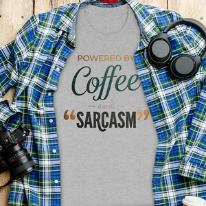 Puede incluir: Una camiseta gris con el texto POWERED BY Coffee and SARCASM en letras marrones, verdes y negras. La camiseta está debajo de una camisa de cuadros azul, verde y blanca, acompañada de auriculares negros y una cámara.