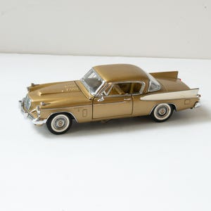 May include: A gold-colored vintage model car with white-walled tires and chrome accents. The classic car has a sleek design with a long hood and a distinctive fin on the rear. The car is a collectible item.