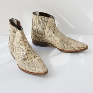 May include: A pair of beige and brown snake-skin patterned ankle boots. The boots have a pointed toe and a low heel. The leather boots are displayed on a white surface.