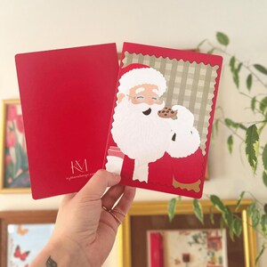 Könnte beinhalten: Eine rote Grußkarte mit einer Illustration vom Weihnachtsmann, der einen Schokoladenkeks isst. Die Karte hat einen gewellten Rand und einen grün-weiß karierten Hintergrund. Die Rückseite ist einfarbig rot mit dem weißen Text KgMarieDesign.com.
