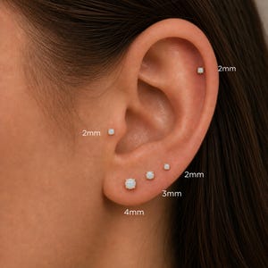 May include: A set of five small round opal stud earrings set in gold metal, displayed in an ear. The studs are labeled with sizes 2mm, 3mm, and 4mm, showing various placements including the helix, tragus, and three lobe positions.