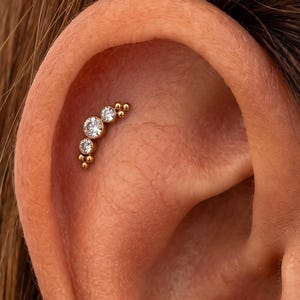 May include: A gold-toned curved cartilage earring featuring three clear round-cut stones of varying sizes arranged in a row, accented by small gold beads at both ends, secured in the upper ear cartilage.