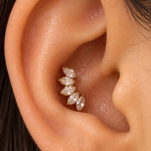 May include: A gold-toned ear cartilage piercing jewelry piece featuring five marquise-cut clear stones arranged in a curved, fan-like cluster. The stones are set in a metal frame that follows the natural contour of the inner ear cartilage.