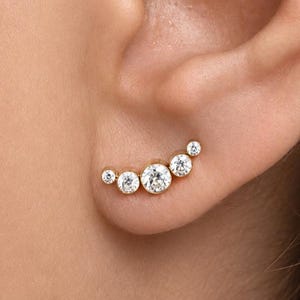 May include: A gold-toned ear climber earring featuring five round, clear, faceted stones arranged in a graduated arc. The stones are set in metal bezels, with the largest stone positioned in the center and smaller stones tapering off toward each end.