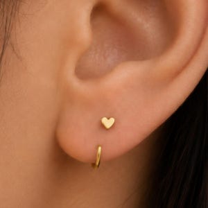 Könnte beinhalten: Ein kleiner goldener Ohrstecker in Herzform, der über einem dünnen goldenen Creolen-Ohrring platziert ist. Beide Schmuckstücke haben eine glatte, polierte Oberfläche und ein schlichtes Design, was einen abgestimmten Look am Ohrläppchen ergibt.