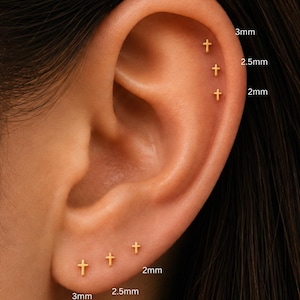 May include: A set of six small gold cross stud earrings arranged on an earlobe and cartilage. The earrings are labeled with sizes of 2mm, 2.5mm, and 3mm, indicating the varying dimensions of the cross designs displayed in this ear piercing set.