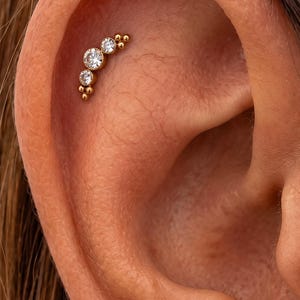 May include: A gold-toned curved ear cartilage stud featuring three clear round-cut stones of varying sizes arranged in a row, accented by small gold spheres at each end, and a separate single round clear stone stud located on the earlobe.