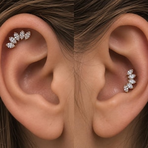 May include: A silver-toned ear cartilage piercing featuring a curved row of five marquise-cut clear stones. The jewelry is shown in two positions: one along the upper rim of the ear and another placed within the conch area of the ear.
