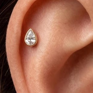Può includere: Un orecchino a lobo in oro con una pietra preziosa trasparente sfaccettata a forma di pera, fissata in una montatura a lunetta liscia. Il gioiello è posizionato nella zona della cartilagine superiore di un orecchio, con una forma a goccia.