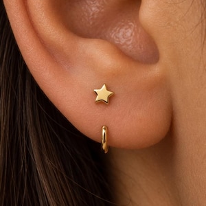May include: A gold star-shaped stud earring positioned above a small, plain gold hoop earring. Both pieces of jewelry feature a polished, reflective metallic finish and are worn in the earlobe, showcasing a simple and clean design aesthetic.