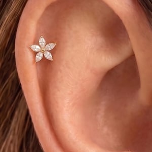 Puede incluir: Un pendiente de botón dorado con diseño de flor de cinco pétalos. Cada pétalo está formado por una piedra preciosa transparente de talla marquesa, y el centro de la flor está acentuado con una pequeña cuenta dorada, redonda y pulida.