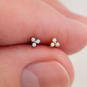 May include: Two small stud earrings, one in silver-toned metal and one in gold-toned metal. Each earring features a cluster of three clear, round-cut stones arranged in a triangular shape, held by small metal prongs on a flat base.