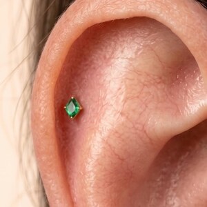 May include: A small gold stud earring featuring a kite-shaped green gemstone. The jewelry is positioned in the upper cartilage area of an ear. The gemstone is faceted and held in place by a gold setting with four small prongs at the corners.