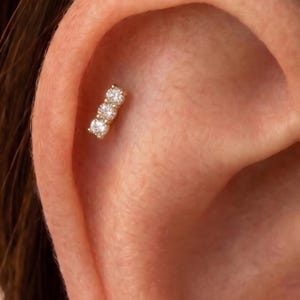 May include: A gold ear piercing stud featuring three small, round, clear, faceted stones arranged in a vertical line. The jewelry is set against the skin of an ear, showcasing a polished metal setting that holds each stone securely in place.
