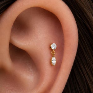 May include: A gold-toned earring featuring a small, round, clear gemstone stud connected to a dangling, marquise-shaped clear gemstone. The jewelry piece is secured in the cartilage area of an ear, showcasing a polished metal finish and two faceted stones.
