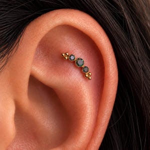 May include: A gold curved cartilage earring featuring three round, dark teal-colored gemstones set in a row. The piece is accented with small gold spheres at both ends of the curved bar, creating a delicate, linear design worn in the upper ear.
