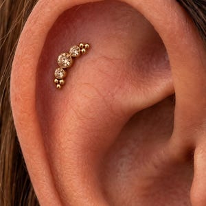 May include: A gold-toned curved cartilage earring featuring three round, light brown faceted stones set in a row, accented by small gold spheres at both ends of the piece. The jewelry is positioned along the upper curve of the ear cartilage.