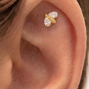 May include: A gold-toned bee-shaped ear stud featuring two clear, pear-shaped faceted stones as wings and a small, ribbed gold-toned body. The jewelry is positioned in the upper cartilage area of an ear, showcasing a detailed insect design.