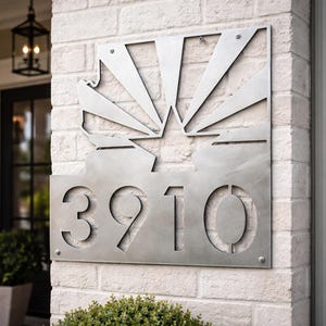 May include: A silver metal address sign with the numbers "3910" cut out. The sign features a stylized sunburst design above the numbers and is mounted on a white brick wall. The sign is attached with four screws.