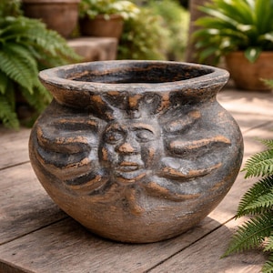 Puede incluir: Una maceta redonda de terracota desgastada con un diseño de cara de sol. La maceta tiene una abertura ancha y un borde oscuro. La cara del sol está en relieve, con una expresión serena. La maceta está sobre una superficie de madera, con vegetación en el fondo.