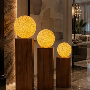 Puede incluir: Tres lámparas esféricas decorativas con superficie texturizada, cada una iluminada con un brillo cálido. Las lámparas se colocan sobre pedestales individuales de madera oscura de diferentes alturas, creando una exhibición visualmente atractiva. El entorno es un espacio interior.