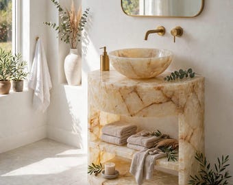 May include: A semi-circular bathroom vanity crafted from translucent, cream-coloured stone with a matching bowl-shaped vessel sink. The unit features two open shelves holding folded beige towels and a small candle, complemented by gold-toned hardware.