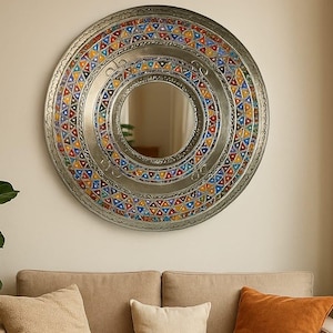 Puede incluir: Un espejo redondo de color plateado con un diseño geométrico colorido. El espejo está colgado en una pared sobre un sofá beige con almohadas y una manta.