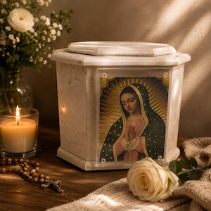 Puede incluir: Una urna funeraria de piedra beige con forma octogonal y tapa extraíble. El panel frontal presenta un marco de acrílico transparente que muestra una imagen colorida de la Virgen de Guadalupe con halo dorado, manto verde oscuro y túnica rosa.