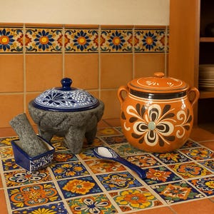 May include: A set of Mexican pottery kitchenware, including a molcajete and tejolote, a covered casserole dish, and a serving spoon. The pottery is decorated with traditional blue and white floral patterns. The items are displayed on a colorful tiled surface.