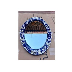 May include: Oval mirror with a blue and white floral design border. The mirror reflects a blue sky and a terracotta roof. The border features a repeating pattern of stylized flowers and leaves, adding a decorative touch to the mirror.
