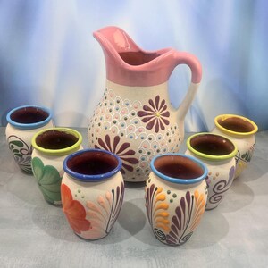 Puede incluir: Un juego de jarra y tazas de cerámica. La jarra es beige con detalles rosas y diseños florales. Las tazas son beige con patrones florales coloridos y bordes de colores. Un juego de utensilios de cocina decorativo y funcional.