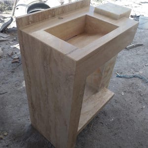 May include: A beige stone bathroom sink with a rectangular basin and a raised backsplash. The sink has a built-in soap dish and a lower shelf. The stone has a natural, veined pattern.