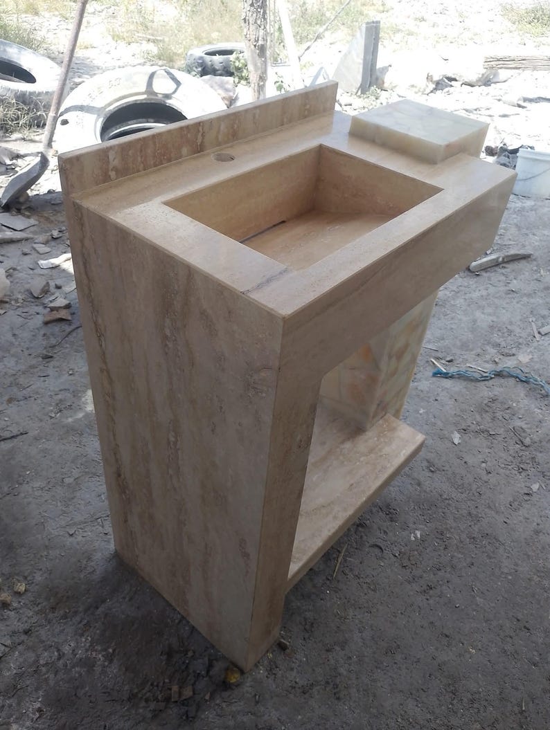 May include: A beige stone bathroom sink with a rectangular basin and a raised backsplash. The sink has a built-in soap dish and a lower shelf. The stone has a natural, veined pattern.