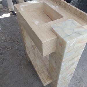 May include: A beige stone bathroom vanity with a built-in rectangular sink. The vanity has a shelf and a decorative column with a mosaic pattern. The overall design is modern and minimalist.