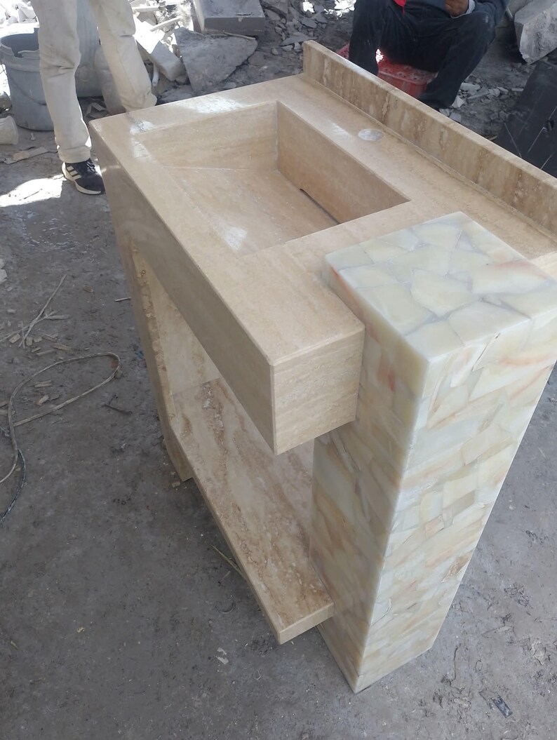 May include: A beige stone bathroom vanity with a built-in rectangular sink. The vanity has a shelf and a decorative column with a mosaic pattern. The overall design is modern and minimalist.