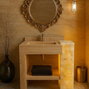 May include: A luxurious bathroom scene featuring a stone vanity with a built-in sink and a gold-framed mirror. A decorative light fixture hangs above. A black vase with branches and a gold trash can complete the elegant design.