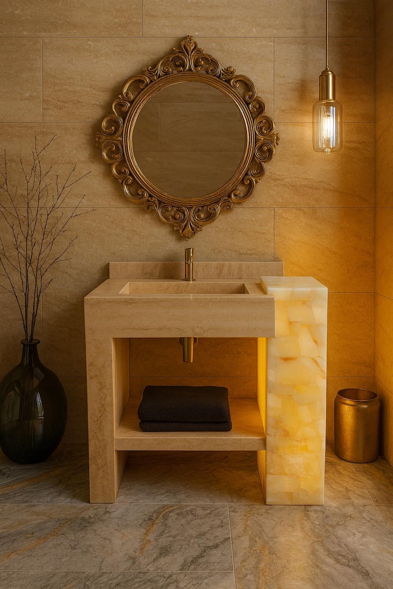 May include: A luxurious bathroom scene featuring a stone vanity with a built-in sink and a gold-framed mirror. A decorative light fixture hangs above. A black vase with branches and a gold trash can complete the elegant design.