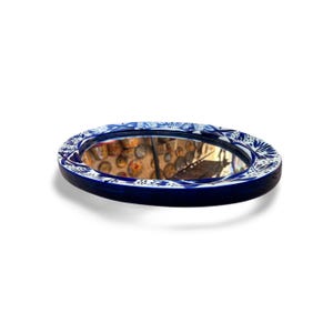 May include: Oval-shaped mirror with a dark blue frame and a white floral pattern. The mirror reflects a room with decorative items. The frame is approximately 10 inches in diameter, adding a decorative touch to any space.