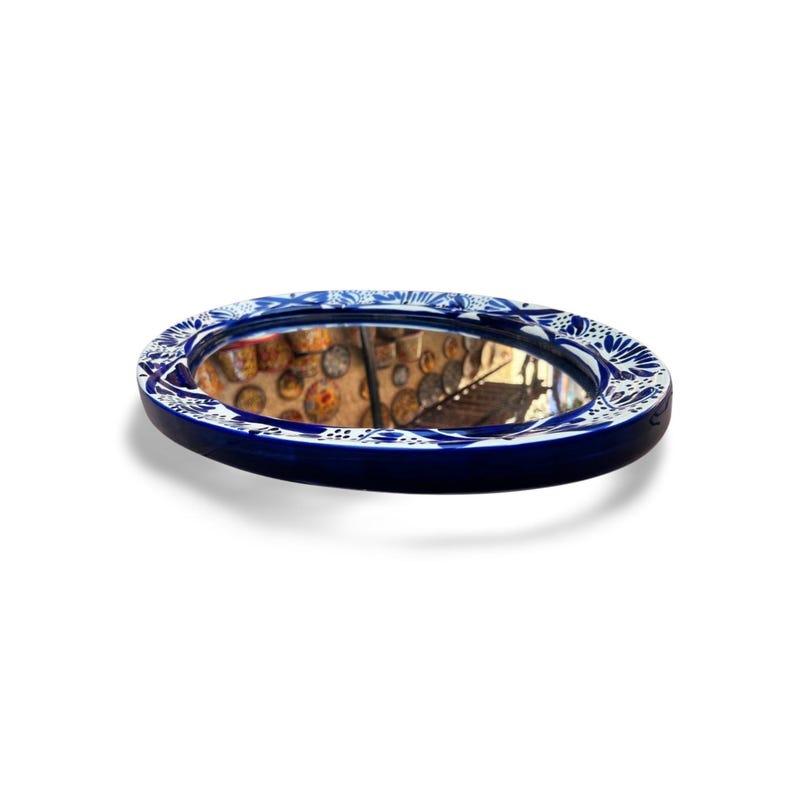 May include: Oval-shaped mirror with a dark blue frame and a white floral pattern. The mirror reflects a room with decorative items. The frame is approximately 10 inches in diameter, adding a decorative touch to any space.