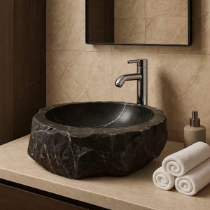 Puede incluir: Un lavabo de piedra negra con un exterior texturizado y un interior liso. Un grifo cromado está montado sobre el lavabo. Toallas blancas enrolladas y un dispensador de jabón están sobre la encimera. Un accesorio de baño único.