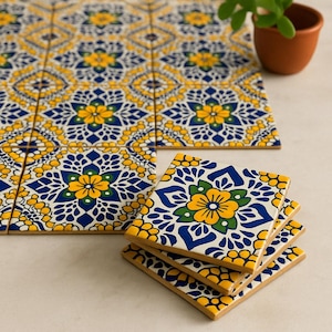 Puede incluir: Un conjunto de baldosas y posavasos cuadrados de cerámica con un diseño floral. Las baldosas y los posavasos presentan un patrón de flores amarillas, hojas azules y detalles blancos sobre un fondo crema. Los posavasos están apilados.