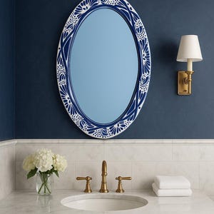 Puede incluir: Espejo ovalado con un borde de diseño floral azul y blanco, montado en una pared azul marino. El tocador del baño presenta un lavabo blanco, un grifo dorado y un pequeño ramo de flores blancas en un jarrón de cristal.