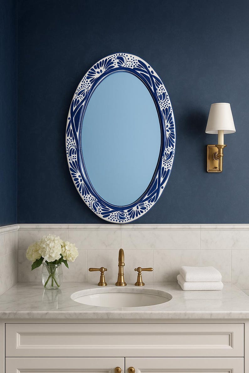 May include: Oval mirror with a blue and white floral design border, mounted on a navy blue wall. The bathroom vanity features a white sink, gold faucet, and a small bouquet of white flowers in a glass vase.