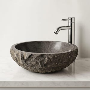 Puede incluir: Un lavabo de piedra gris oscuro con un exterior rugoso y texturizado. El lavabo redondo se asienta sobre una encimera blanca, con un grifo cromado a la derecha. El interior del lavabo es liso y gris oscuro.